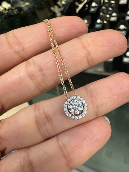 An 18 carat rose gold halo cluster pendant featuring a round brilliant cut diamond center stone with a halo of smaller round diamonds, suspended on a slender gold chain. on hand
