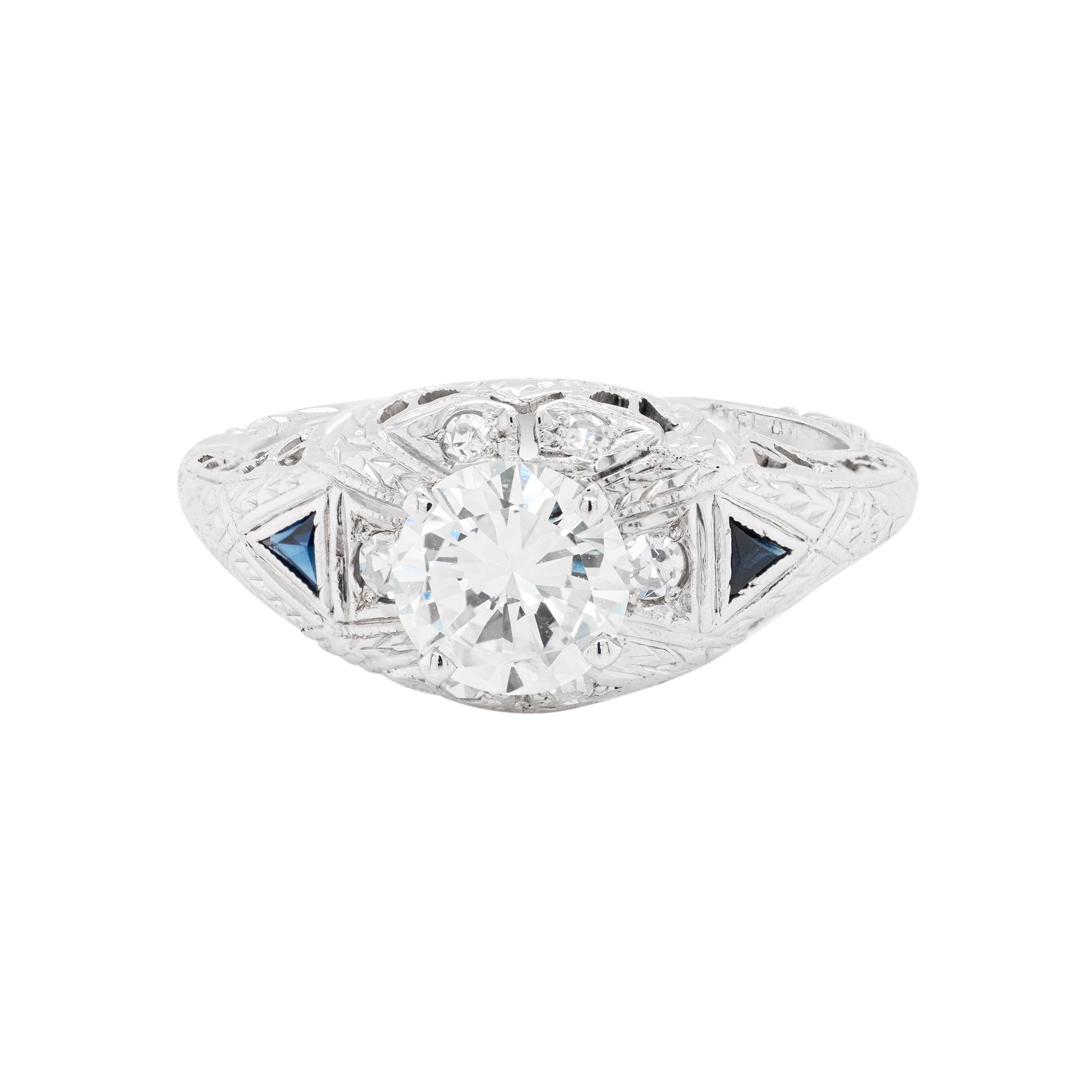 A hand-carved platinum engagement ring with a round brilliant cut diamond centerpiece, accompanied by two triangle-shaped sapphires and six grain-set diamonds.