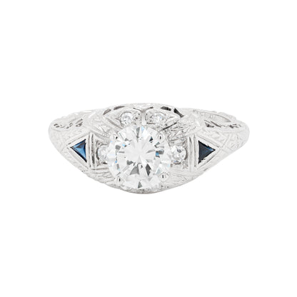 A hand-carved platinum engagement ring with a round brilliant cut diamond centerpiece, accompanied by two triangle-shaped sapphires and six grain-set diamonds.