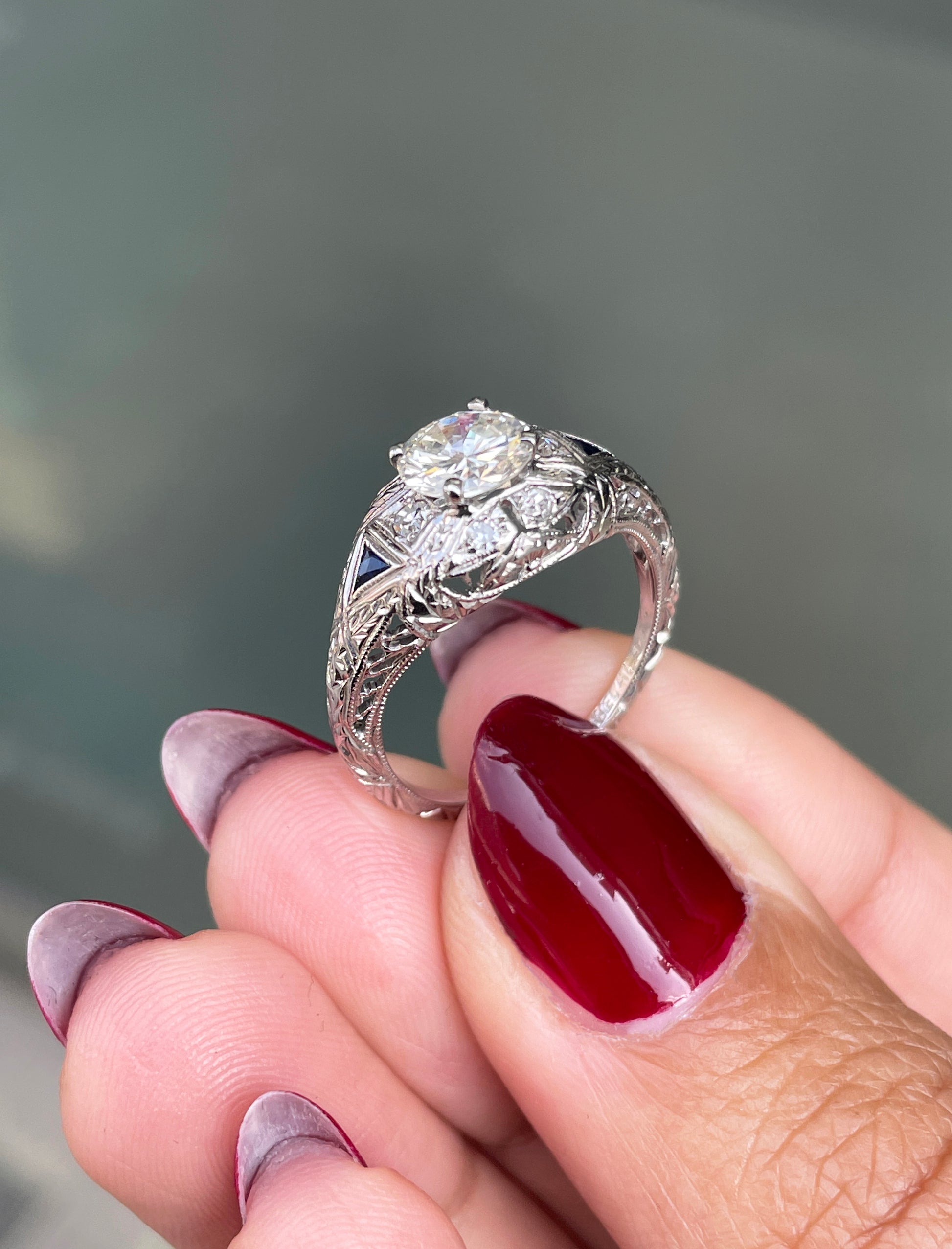 A hand-carved platinum engagement ring with a round brilliant cut diamond centerpiece, accompanied by two triangle-shaped sapphires and six grain-set diamonds on hand