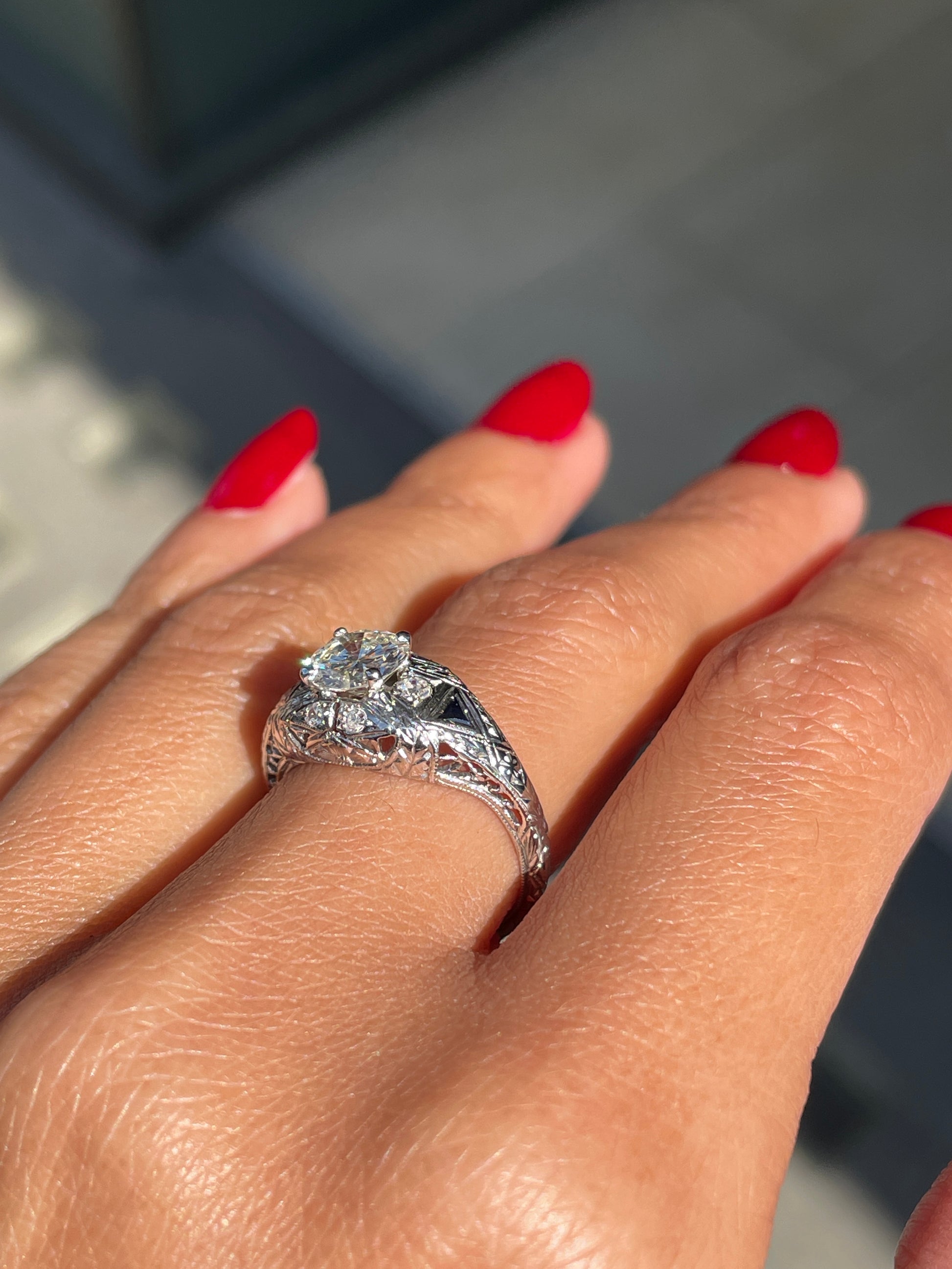 A hand-carved platinum engagement ring with a round brilliant cut diamond centerpiece, accompanied by two triangle-shaped sapphires and six grain-set diamonds on hand