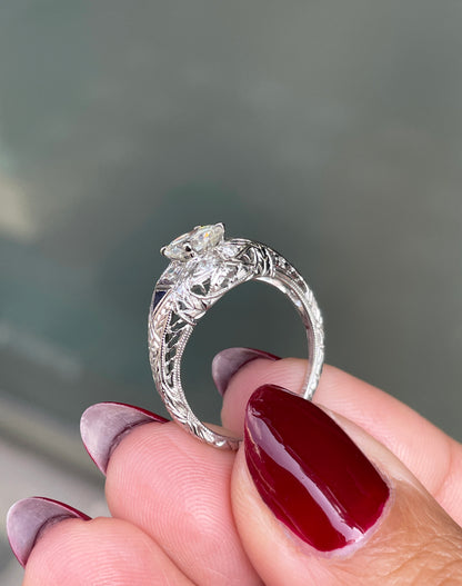 A hand-carved platinum engagement ring with a round brilliant cut diamond centerpiece, accompanied by two triangle-shaped sapphires and six grain-set diamonds on hand
