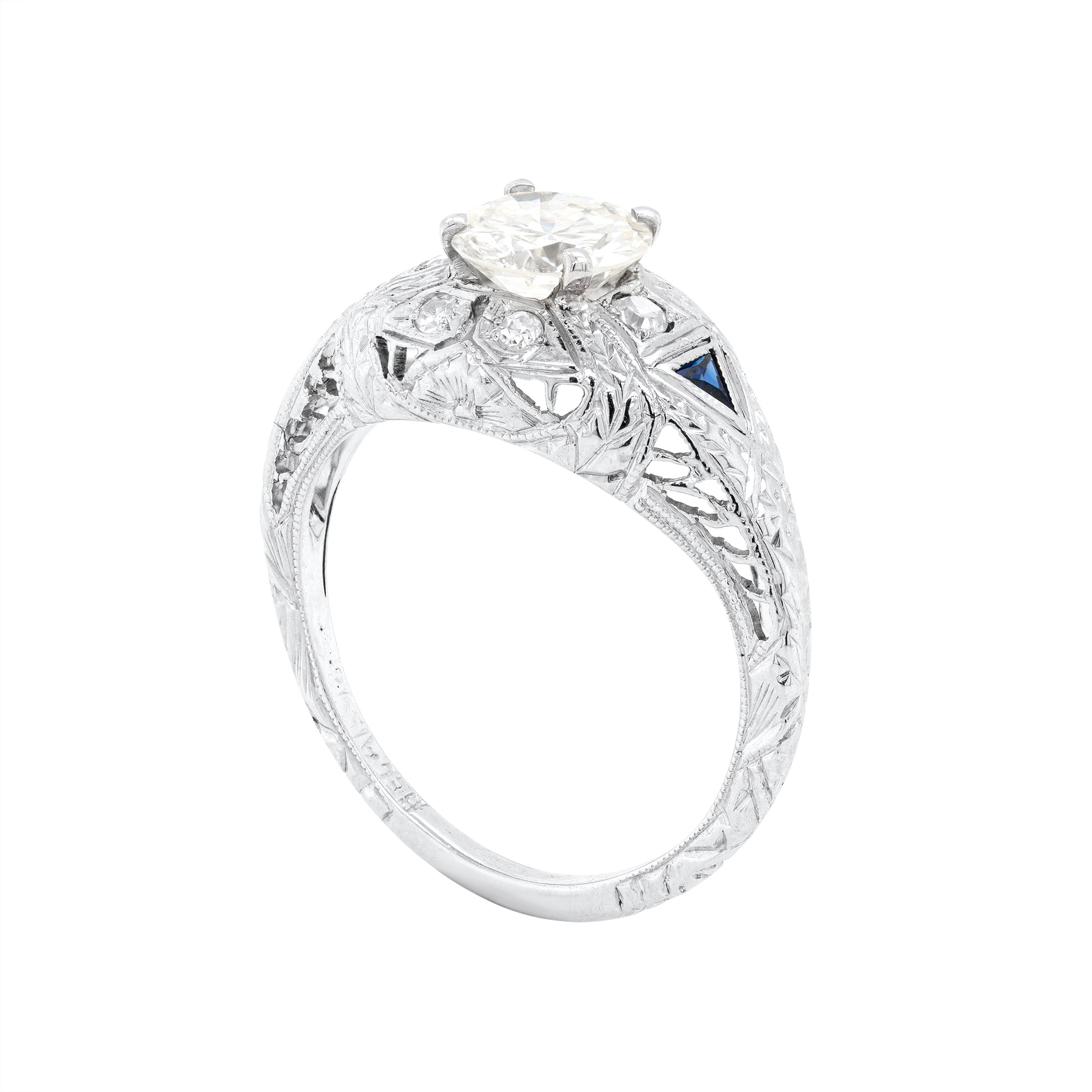 A hand-carved platinum engagement ring with a round brilliant cut diamond centerpiece, accompanied by two triangle-shaped sapphires and six grain-set diamonds 