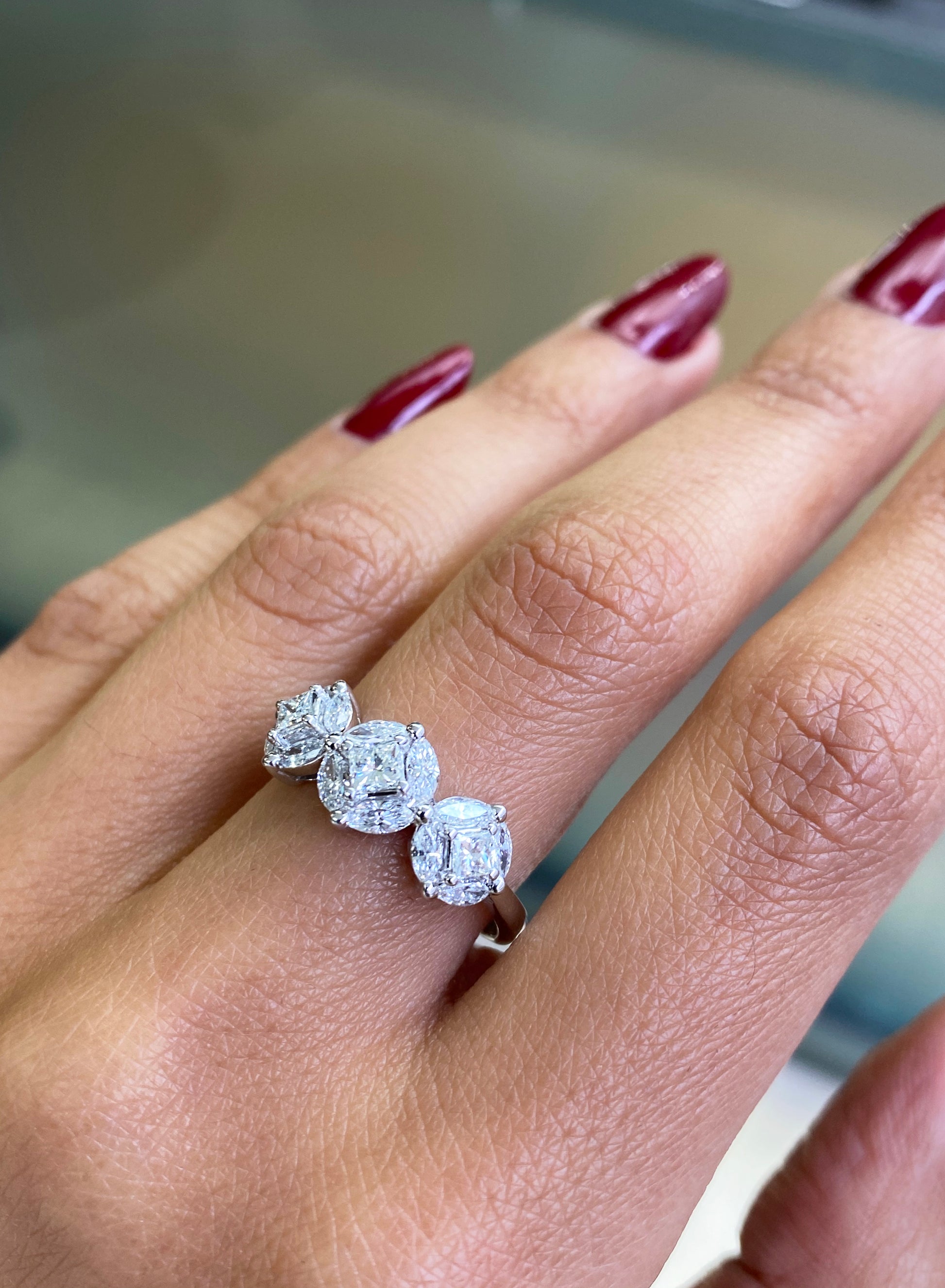 An 18 carat white gold trilogy diamond cluster engagement ring with three circular diamond clusters and a princess cut diamond in the center, set in a white gold band on hand