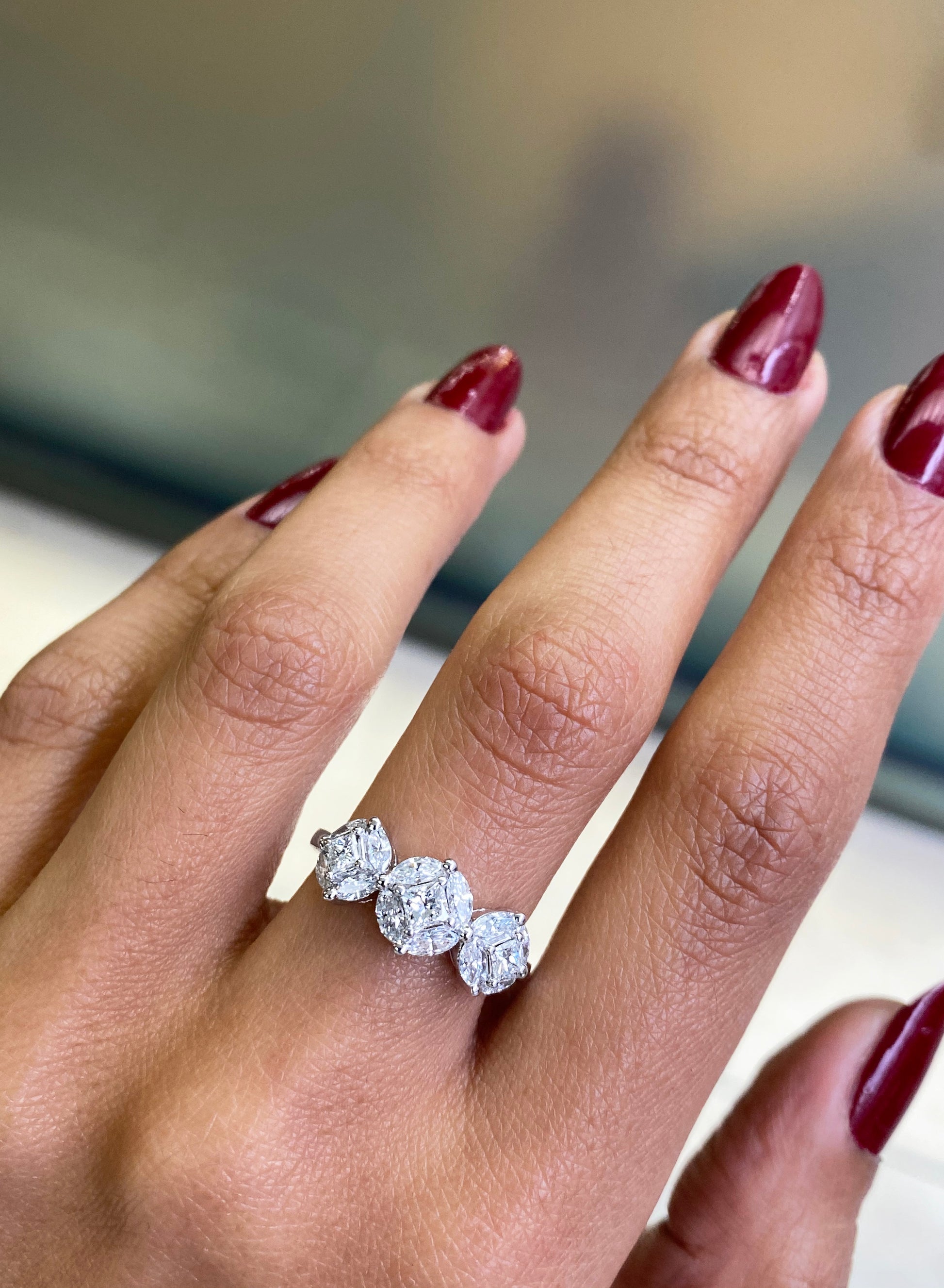 An 18 carat white gold trilogy diamond cluster engagement ring with three circular diamond clusters and a princess cut diamond in the center, set in a white gold band on hand on hand