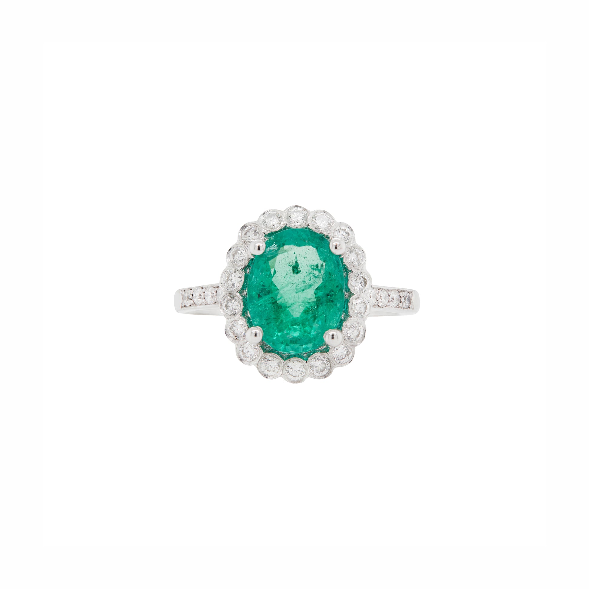 An 18 carat white gold cluster engagement ring with a central oval emerald and a halo of round brilliant cut diamonds.