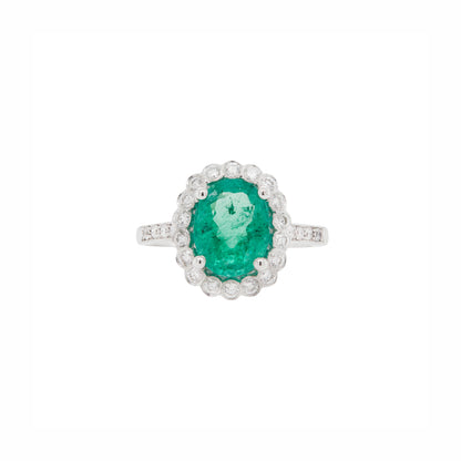 An 18 carat white gold cluster engagement ring with a central oval emerald and a halo of round brilliant cut diamonds.