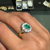 Gold ring with a green gemstone on a finger, with blurred jewelry boxes in the background.