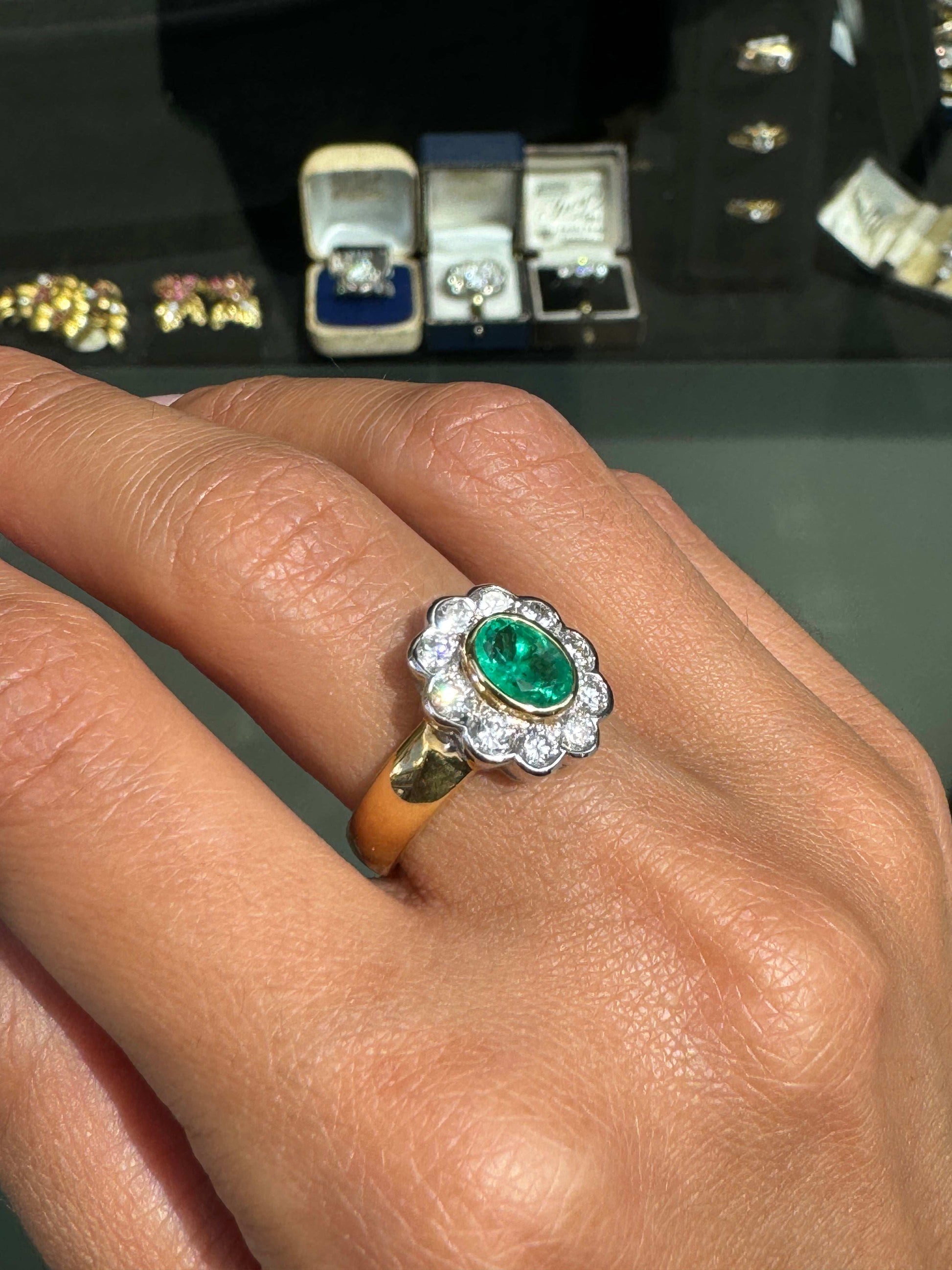 Gold ring with a green gemstone on a finger, with blurred jewelry boxes in the background.