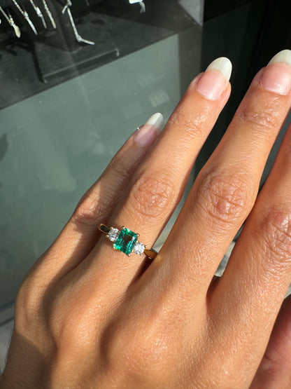 Hand wearing a green emerald and diamond ring on a reflective surface