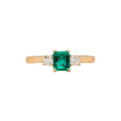 An 18K yellow gold three stone engagement ring featuring a rectangular emerald cut Colombian emerald center stone flanked by two round brilliant cut diamonds.