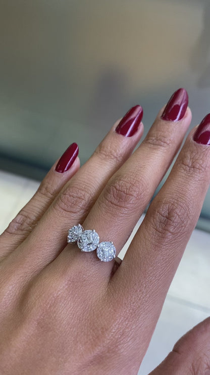 An 18 carat white gold trilogy diamond cluster engagement ring with three circular diamond clusters and a princess cut diamond in the center, set in a white gold band on hand 