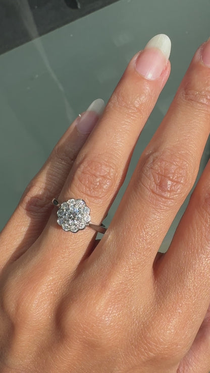 video of A round brilliant cut diamond ring with smaller round brilliant cut diamonds set in a daisy cluster design, mounted in a platinum band.