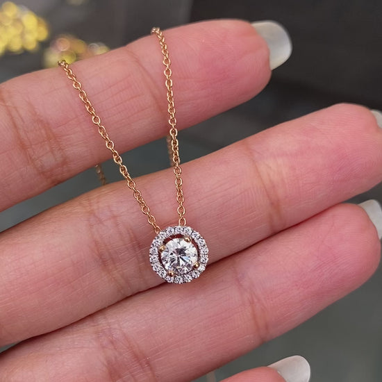 An 18 carat rose gold halo cluster pendant featuring a round brilliant cut diamond center stone with a halo of smaller round diamonds, suspended on a slender gold chain. on hand
