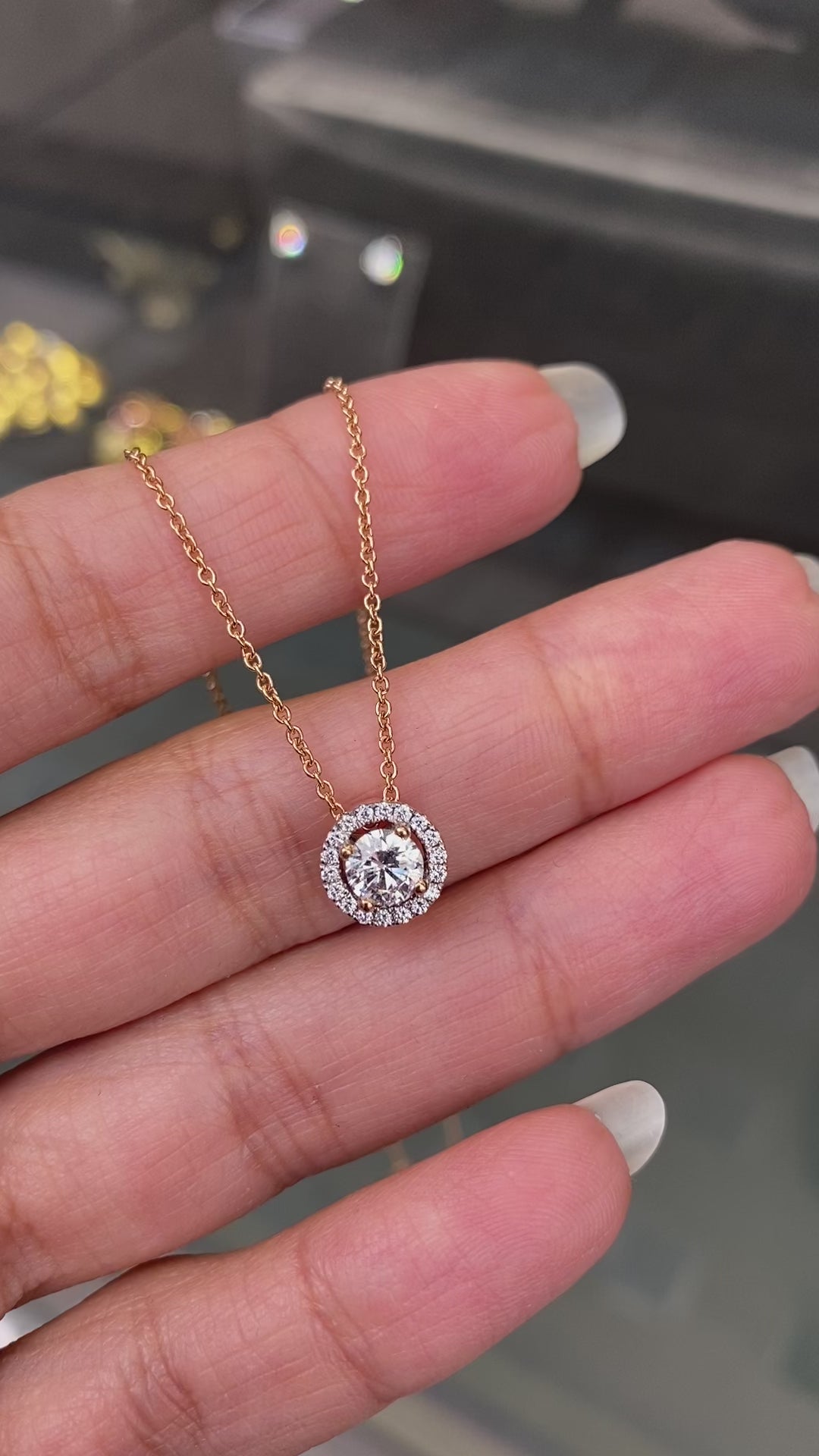 An 18 carat rose gold halo cluster pendant featuring a round brilliant cut diamond center stone with a halo of smaller round diamonds, suspended on a slender gold chain. on hand