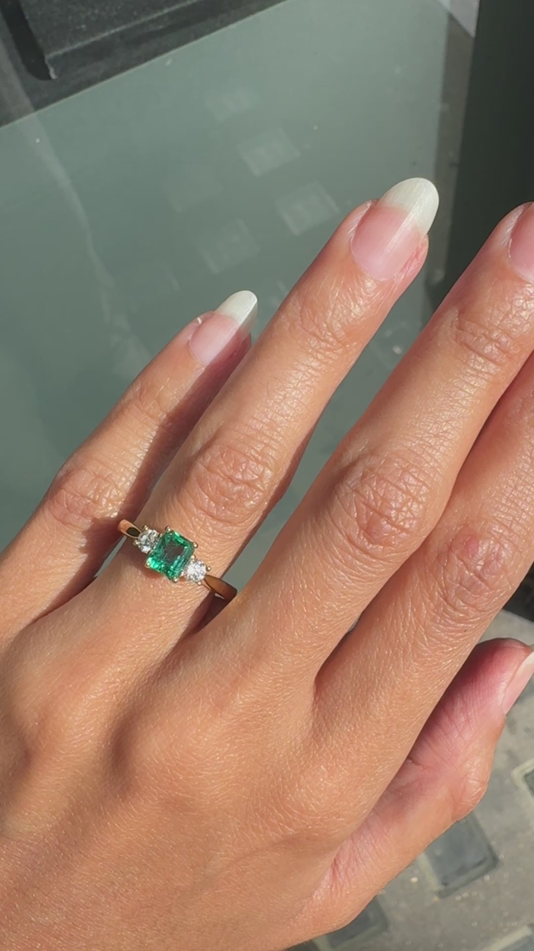 video of An 18K yellow gold three stone engagement ring featuring a rectangular emerald cut Colombian emerald center stone flanked by two round brilliant cut diamonds.